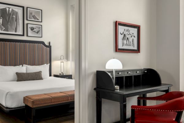 A stylish bedroom with a bed, vintage desk, chairs, and framed art on the walls. A modern lamp sits on the desk.