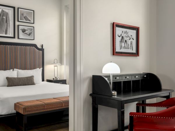 A stylish bedroom with a bed, vintage desk, chairs, and framed art on the walls. A modern lamp sits on the desk.