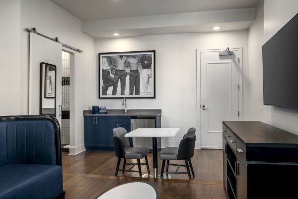 The image shows a modern room with a small table, chairs, a TV, and a wall photo. It has a sleek, minimalistic design with wooden flooring.