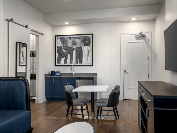 The image shows a modern room with a small table, chairs, a TV, and a wall photo. It has a sleek, minimalistic design with wooden flooring.