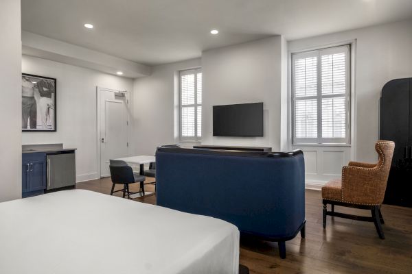 A modern room with a bed, blue couch, TV, and chairs, featuring natural light from large windows and a minimalist design.