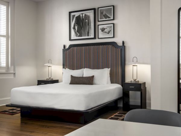 A modern bedroom with a large bed, two side tables, and lamps. Black-and-white photos above the bed and a tall mirror on the right.