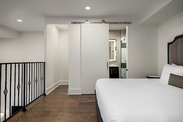 The image shows a modern bedroom with a white bed, wooden floor, sliding barn door, railings, and view into a bathroom area.
