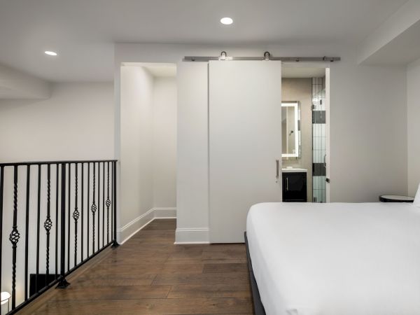The image shows a modern bedroom with a white bed, wooden floor, sliding barn door, railings, and view into a bathroom area.