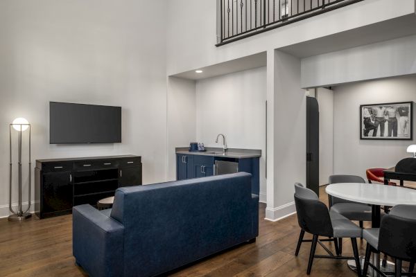 The image shows a modern living room with a sofa, wall-mounted TV, kitchenette, and dining area. There's also artwork and staircase in view.