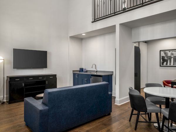 The image shows a modern living room with a sofa, wall-mounted TV, kitchenette, and dining area. There's also artwork and staircase in view.