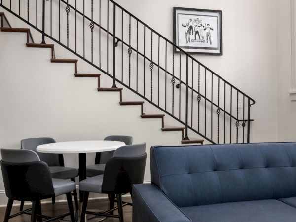 A modern interior with a blue sofa, round table, chairs, staircase, and framed artwork on the wall.