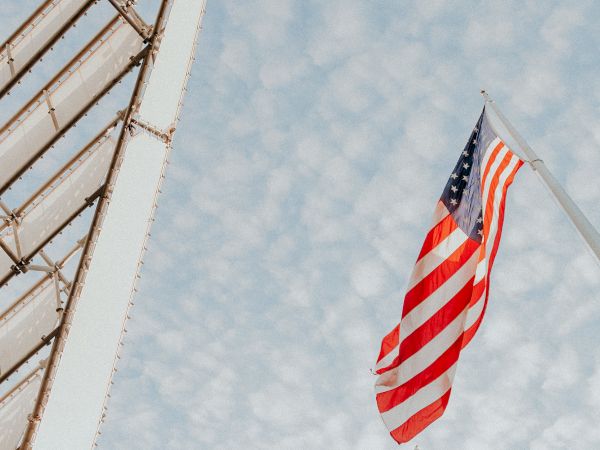 An American flag waves against a sky with scattered clouds, alongside part of a metal structure on the left.