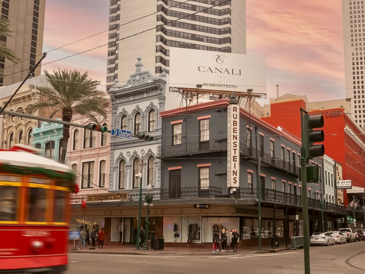A city street with a vintage red trolley, historic buildings, and palm trees against a backdrop of modern skyscrapers under a sunset sky.