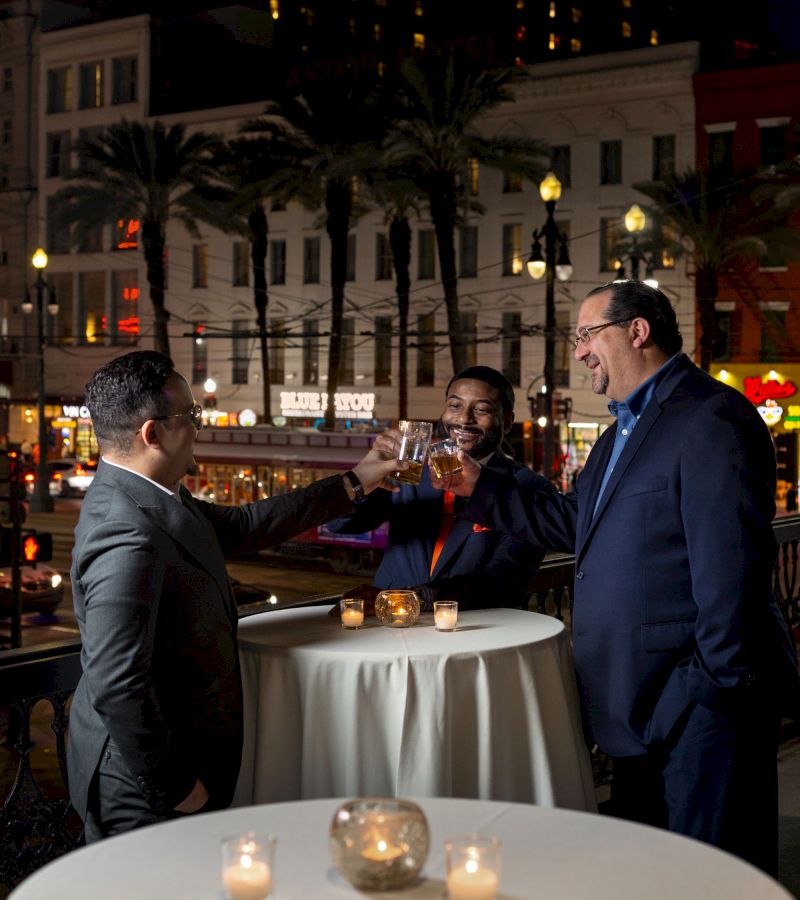 Three men in suits clinking glasses at a table with candles on a balcony overlooking a city street at night.