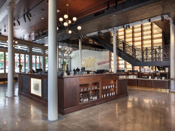 A spacious interior with a wooden bar, ornate staircase, and bottles displayed behind. Brightly lit with large windows and elegant chandeliers.