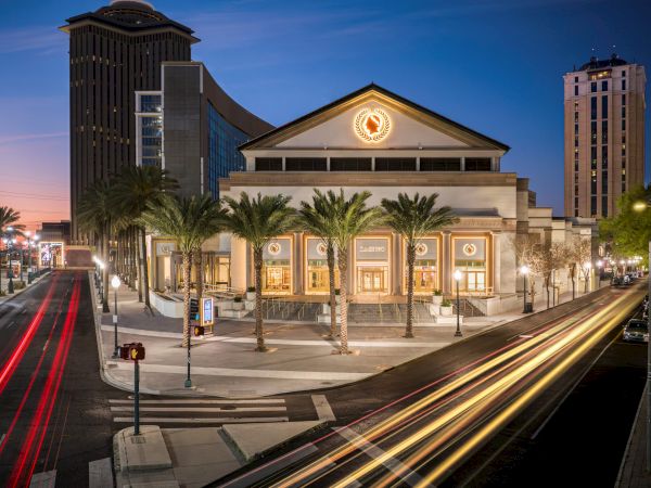 A cityscape at dusk features a grand building with palm trees, light trails from passing traffic, and modern architecture.