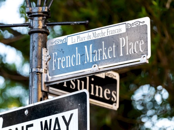 A street sign for "French Market Place" and another partially visible one, with a "One Way" sign below, surrounded by trees.