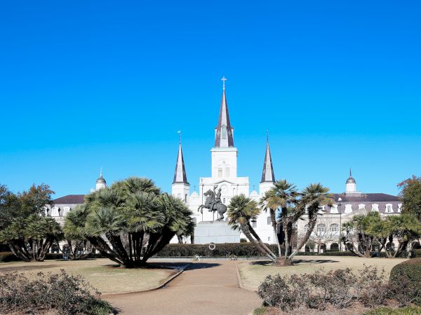 The image shows Jackson Square in New Orleans, featuring the iconic St. Louis Cathedral and an equestrian statue under a clear blue sky.