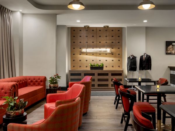 A modern lounge with red seating, tables, plants, wall art, and two suits on display. Ceiling lights provide soft illumination.