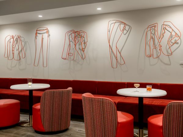 A modern lounge with red seating, white round tables, and artistic metal sculptures resembling clothing on the wall for decoration.