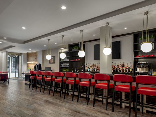 A modern bar with red stools, shelves of bottles, pendant lights, and a wood floor. Cozy seating area visible in the background.