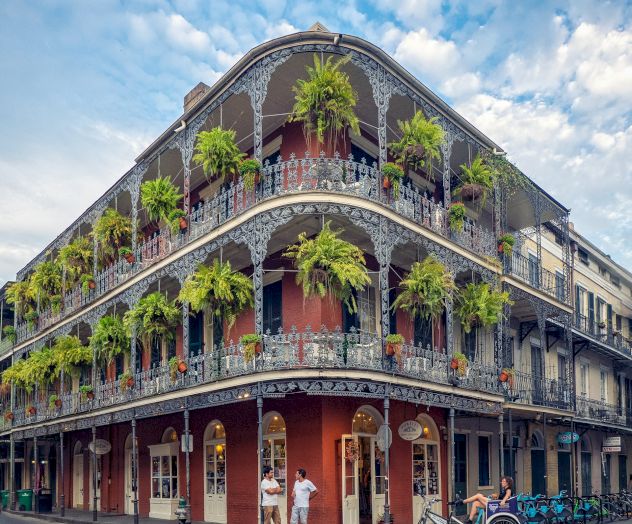 A corner building with ornate ironwork balconies and hanging plants, located on a bustling street corner under a partly cloudy sky.