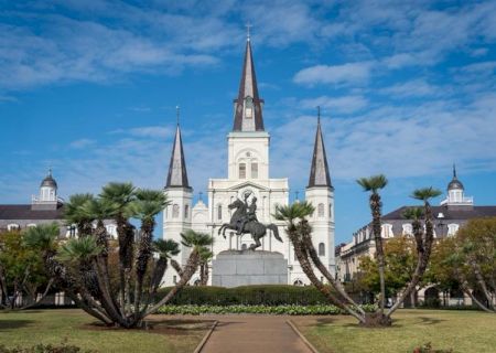 A cathedral with three spires, a statue in the foreground, and palm trees, set against a blue sky with scattered clouds.