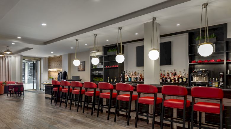 A modern bar with red chairs, hanging lights, and a stocked counter, offering a cozy atmosphere and stylish design.