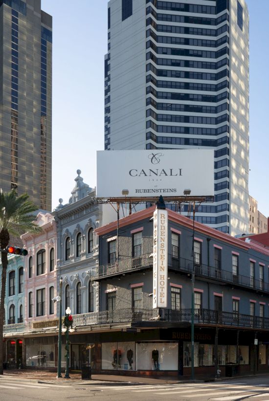 An urban street corner with tall buildings, historic architecture, palm trees, and a billboard above the structures.