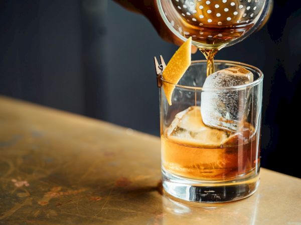 A cocktail being poured into a glass with a large ice cube and garnished with a twist of citrus peel on a bar counter.