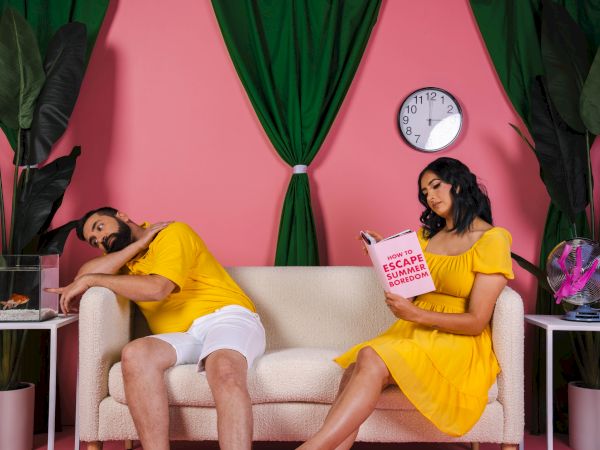 A man sleeps on a sofa while a woman in yellow reads a book. The room is pink with green accents and decorative plants.