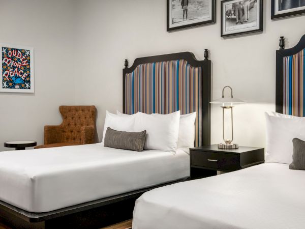 The image shows a hotel room with two beds, striped headboards, a small lamp, framed pictures, and an upholstered chair.