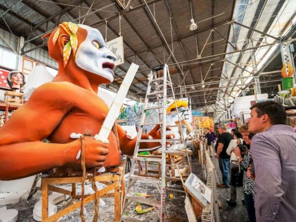 A workshop scene with a large orange figure wearing a white mask, holding a paddle, while people observe and other craftsmen work on sculptural props.
