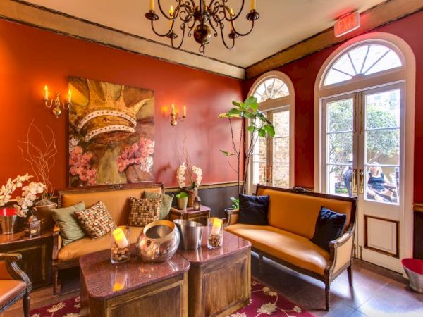 A cozy, elegant lounge with red walls, chandeliers, plush seating, potted plants, and arched French doors letting in light.