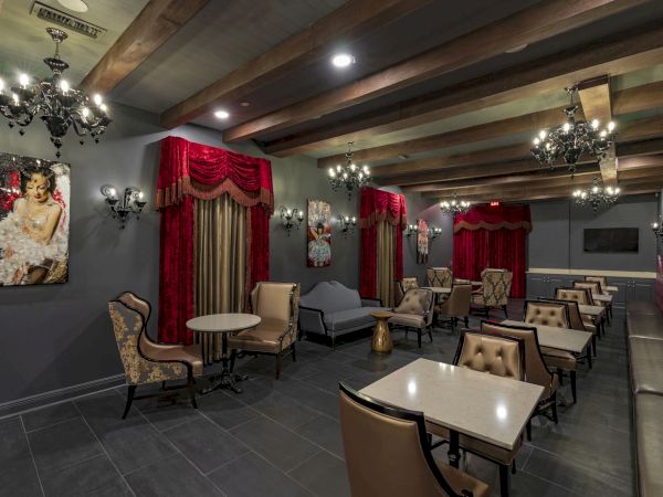 A stylish lounge or cafe with dark walls, red draped curtains, chandeliers, and cozy seating arranged around small tables, offering a luxe vibe.