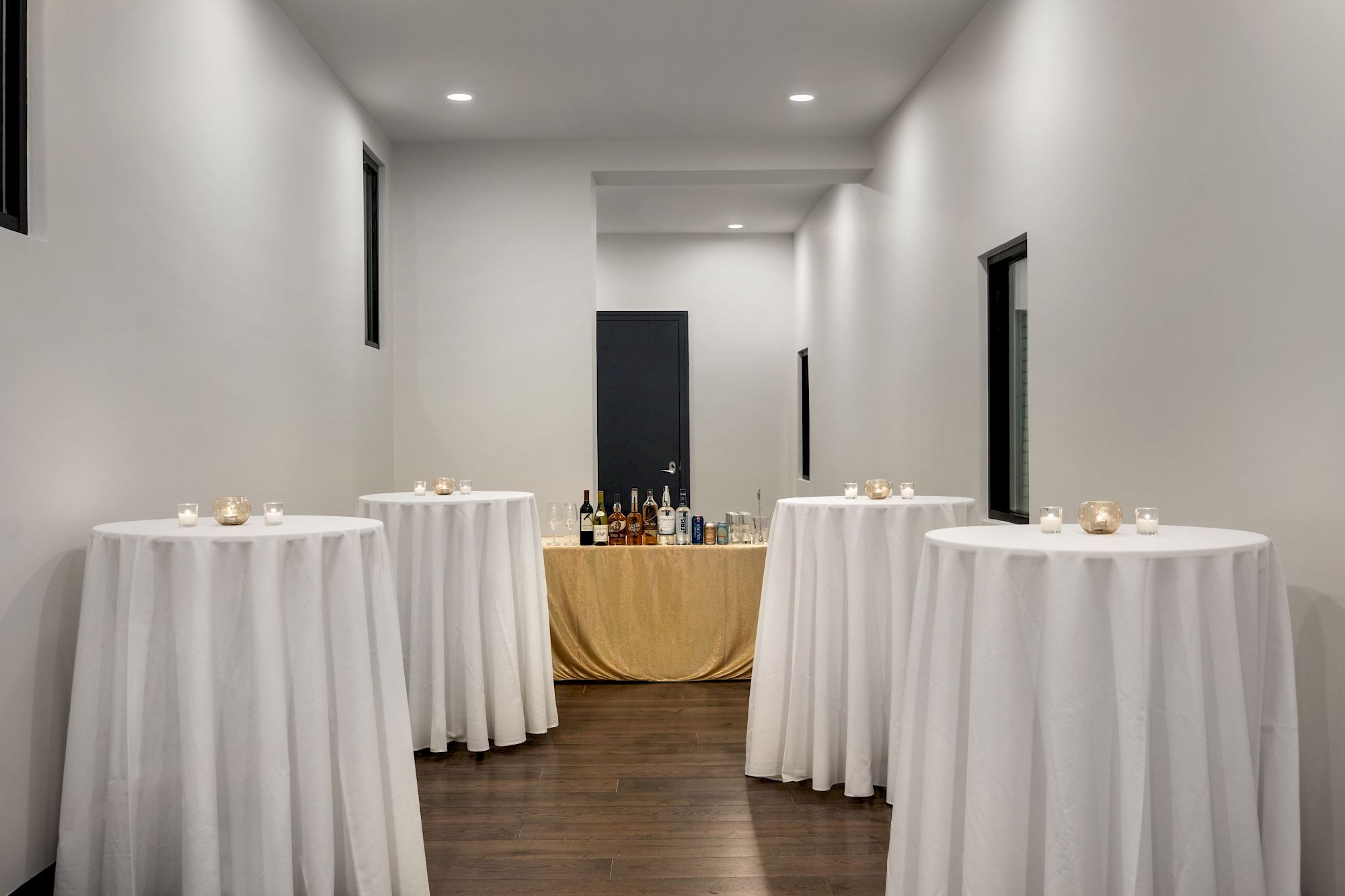 A minimalist event space with white-clothed high-top tables, candles, and a bar setup at the far end in a narrow, well-lit hallway.
