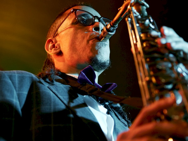 A saxophonist wearing sunglasses and a suit plays under warm stage lights, close-up from below with a microphone in front of his mouth.