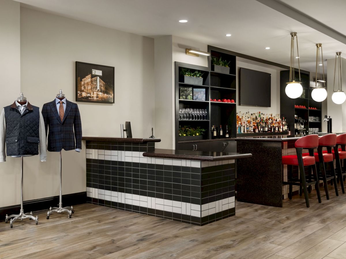 A stylish bar area with black-tiled counter, hanging jackets on stands, wine shelves, and red bar stools under modern pendant lights.