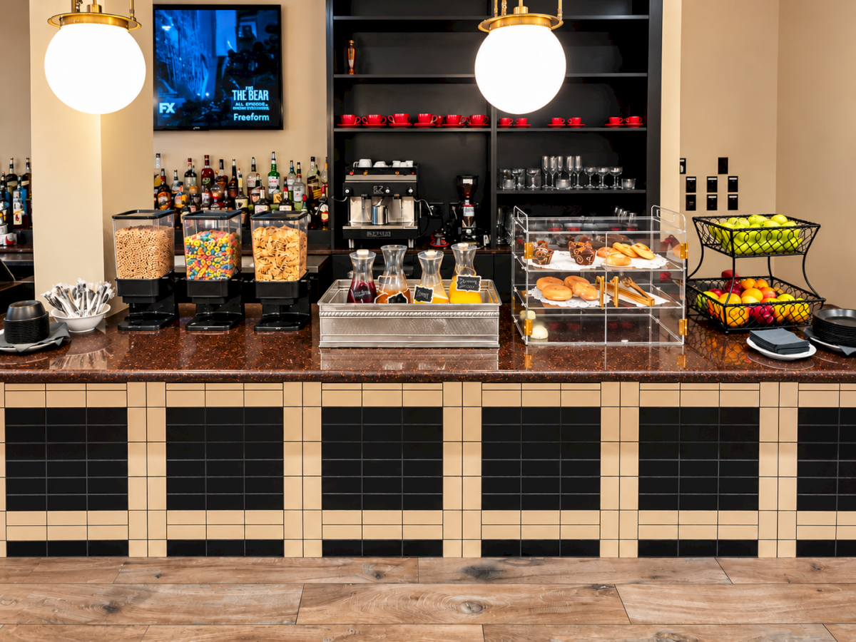 A modern coffee bar with espresso machines, snacks, cups, bottles, and pendant lights behind a tiled counter.