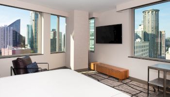 A modern hotel room with large windows, a bed, an armchair, a wall-mounted TV, and views of a city skyline.