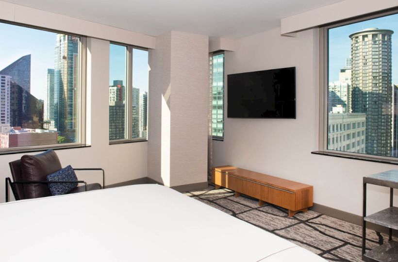 A modern hotel room with large windows, a bed, an armchair, a wall-mounted TV, and views of a city skyline.