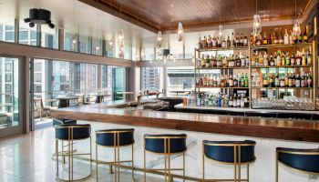 A modern bar with a sleek wooden counter, elevated seating, and a fully stocked shelf against a backdrop of large windows showcasing a cityscape.