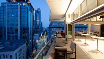 The image shows a modern city view from a high-rise balcony at twilight, with outdoor seating and buildings lining a lit street.