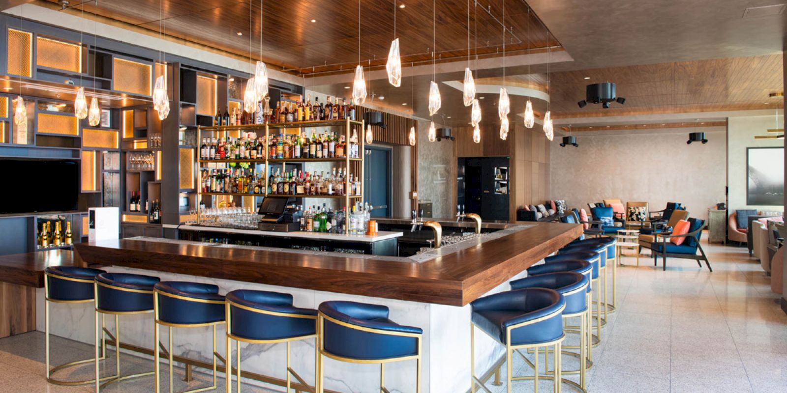 A modern bar with blue bar stools, pendant lights, and a well-stocked bar counter in an open, stylish setting featuring comfy seating.