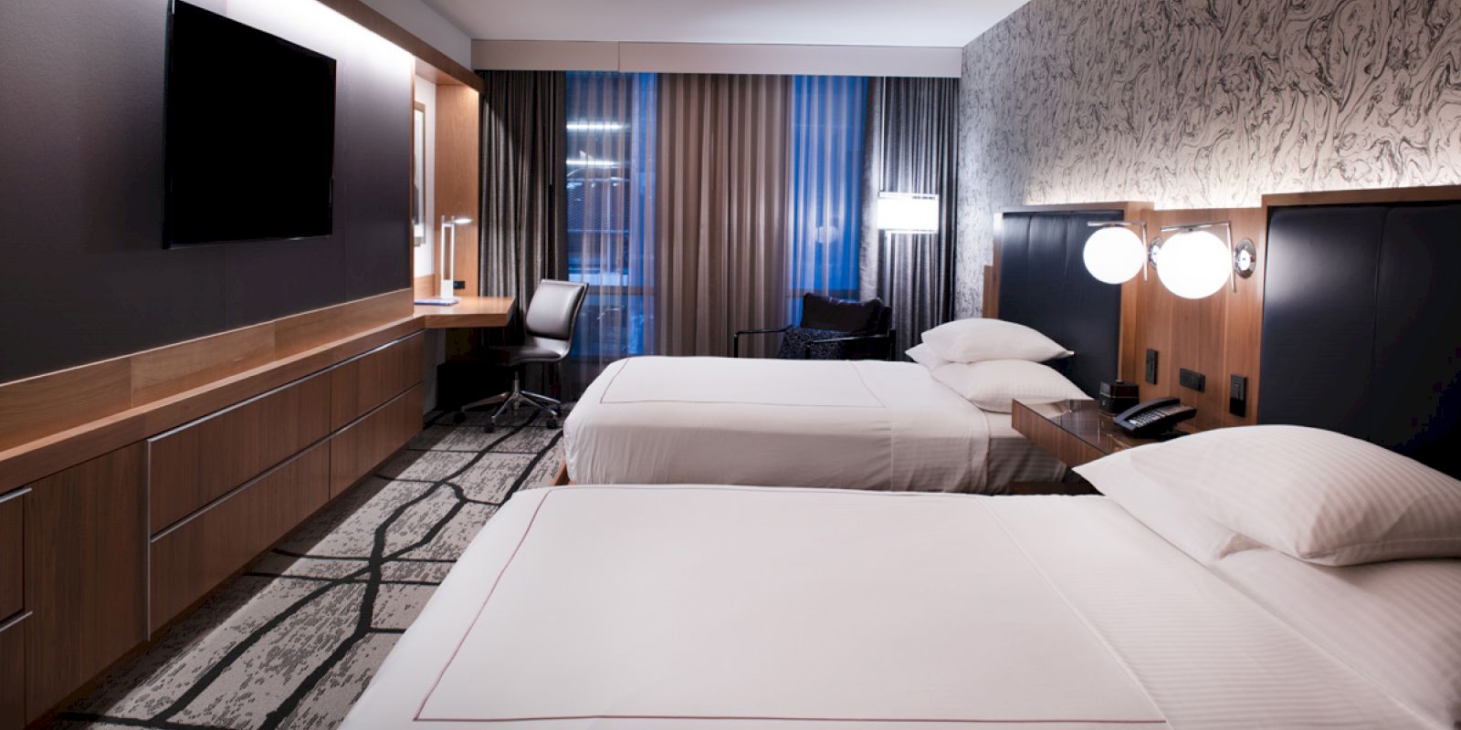 A hotel room with two beds, a TV, desk, chair, and modern decor. The room features a patterned wall, curtains, and ambient lighting.