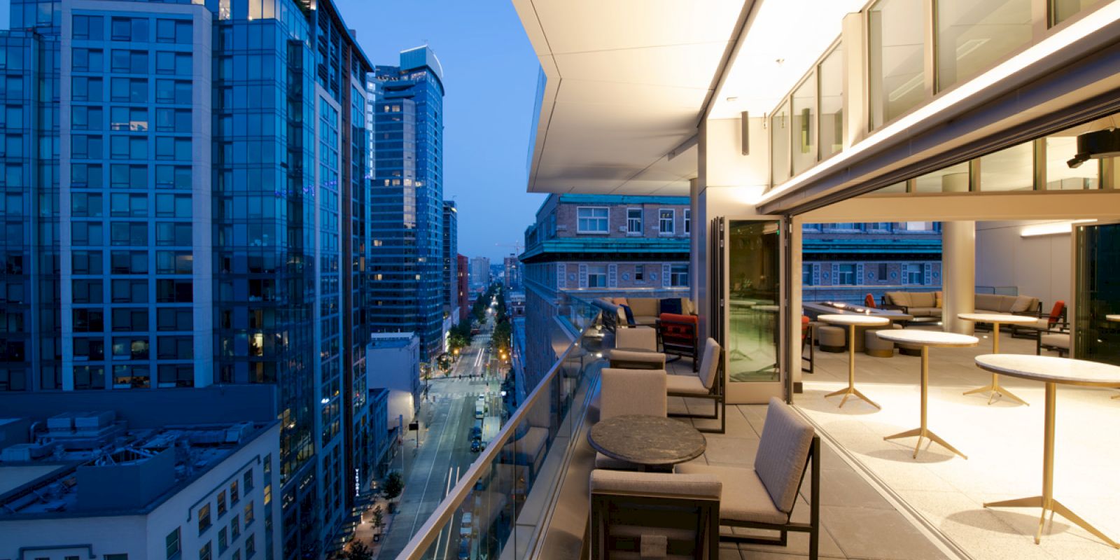 A modern balcony with seating overlooks an urban street and tall buildings, illuminated at twilight, offering a stylish city view.