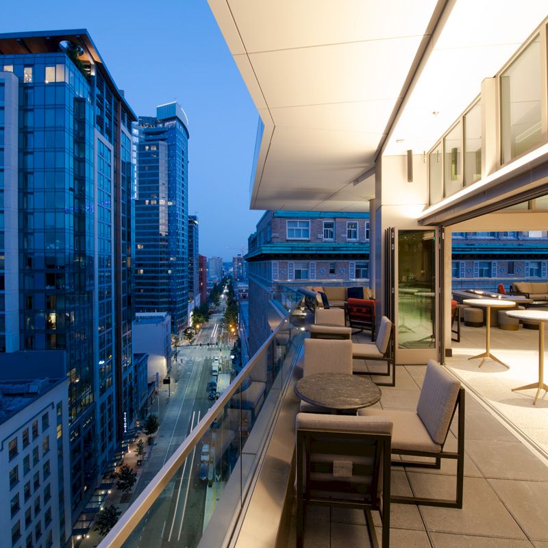 A modern rooftop terrace at dusk with cityscape views, featuring outdoor seating and tables under warm, inviting lights.