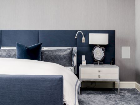 A modern bedroom features a blue upholstered bed, white bedding, and a nightstand with a lamp and decorative stone piece.