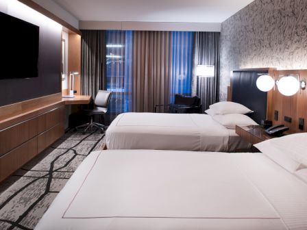 The image shows a modern hotel room with two beds, a TV, a desk with a chair, and lamps. The decor is stylish with patterned wallpaper.