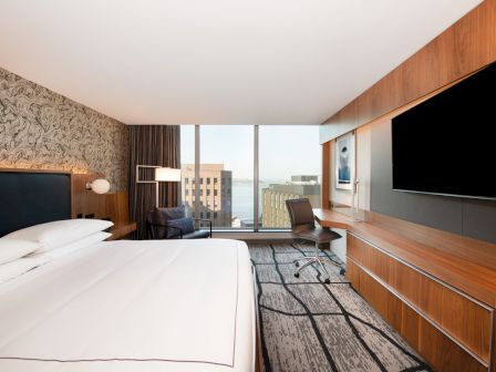 A modern hotel room with a large bed, desk, chair, and flat-screen TV. The window offers a cityscape and water view.