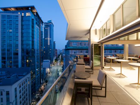 A modern rooftop terrace with seating overlooks a cityscape featuring tall buildings and a street lit up in the evening.