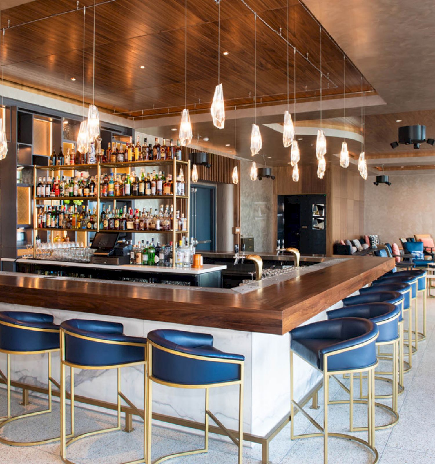 The image shows a modern bar with blue stools, hanging lights, and a well-stocked back shelf. There's seating in the background of the room.