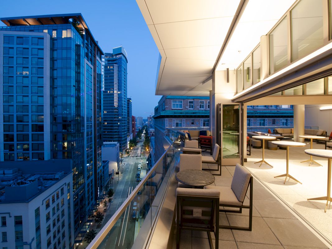 Modern urban balcony overlooking a city street with skyscrapers; features seating, tables, and indoor-outdoor interaction at dusk.