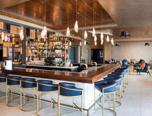 The image shows a modern bar with a wooden counter, blue chairs, hanging lights, and a well-stocked drink selection on shelves.